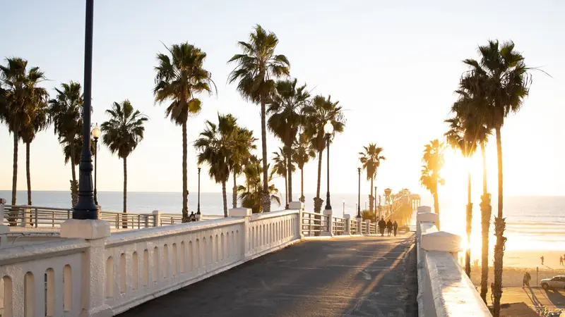 Palm-lined footbridge leading to Oceanside Pier at golden hour, with people walking toward the ocean