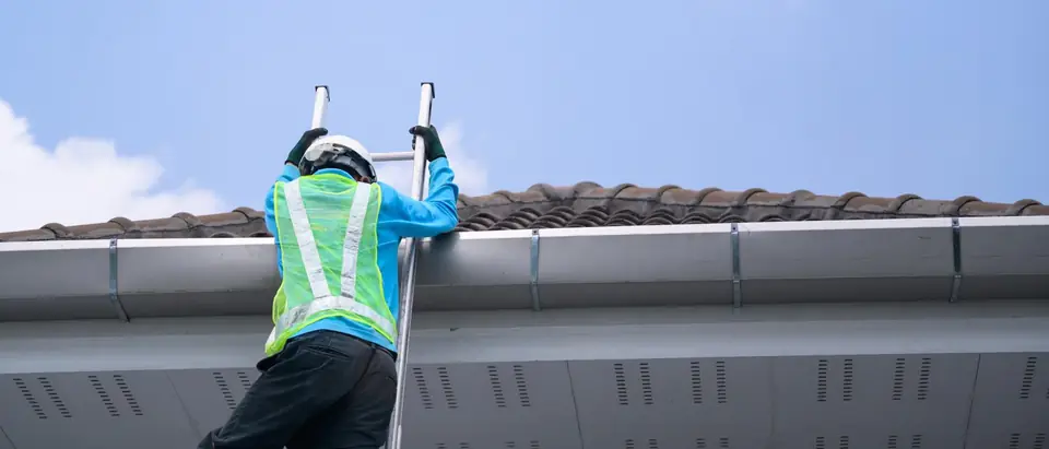 Worker in a hi-vis vest and white hard hat on a ladder at the gutter of a tile roof, blue sky above