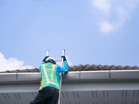 Worker in a hi-vis vest and white hard hat on a ladder at the gutter of a tile roof, blue sky above