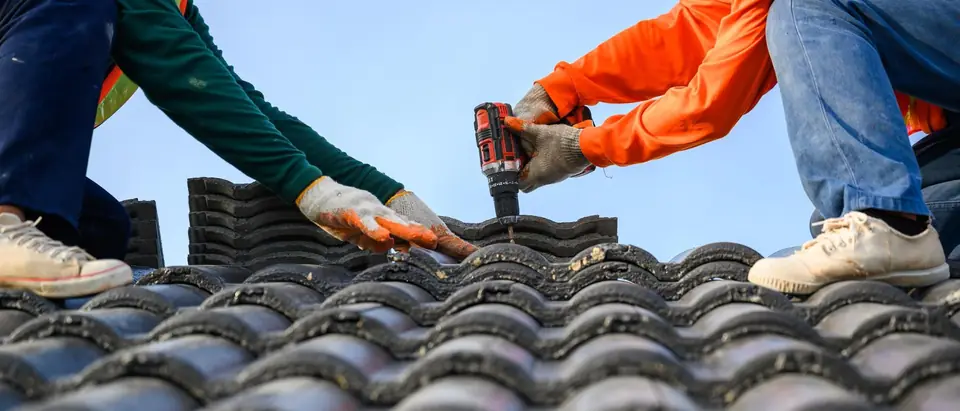 Two roofers in work gloves fasten a concrete roof tile with a cordless drill under a clear blue sky
