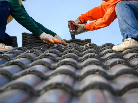 Two roofers in work gloves fasten a concrete roof tile with a cordless drill under a clear blue sky