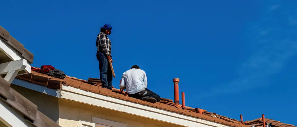 Two workers on the tile roof of a two-story stucco house, loose tiles stacked beside them, deep blue sky