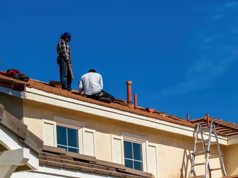 Two workers on the tile roof of a two-story stucco house, loose tiles stacked beside them, deep blue sky